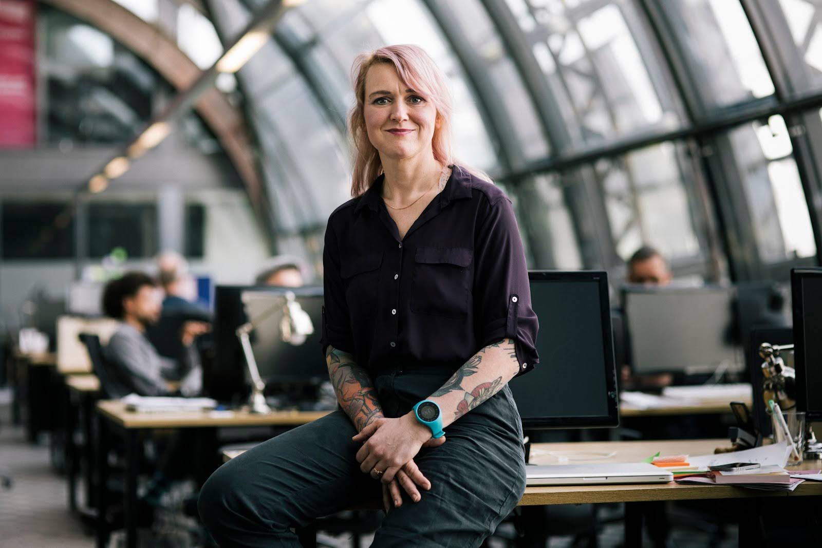 A smiling woman in a modern office space sits facing the camera.