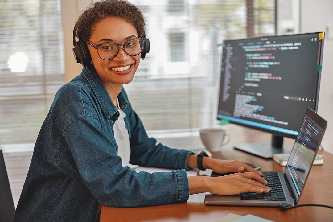 A smiling woman wearing headphones sits at a desk and works on a laptop; an external screen displays computer code.